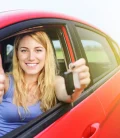 A woman inside a car holding keys with one hand and giving a thumbs up with the other hand, smiling.