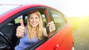 A woman inside a car holding keys with one hand and giving a thumbs up with the other hand, smiling.