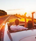 A car on the road at sunset with two people having their hands out, enjoying the view.