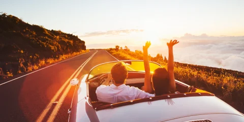 A car on the road at sunset with two people having their hands out, enjoying the view.