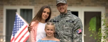 A family of 3 in-front of their house with a proud American flag waving. The father is part of the military.